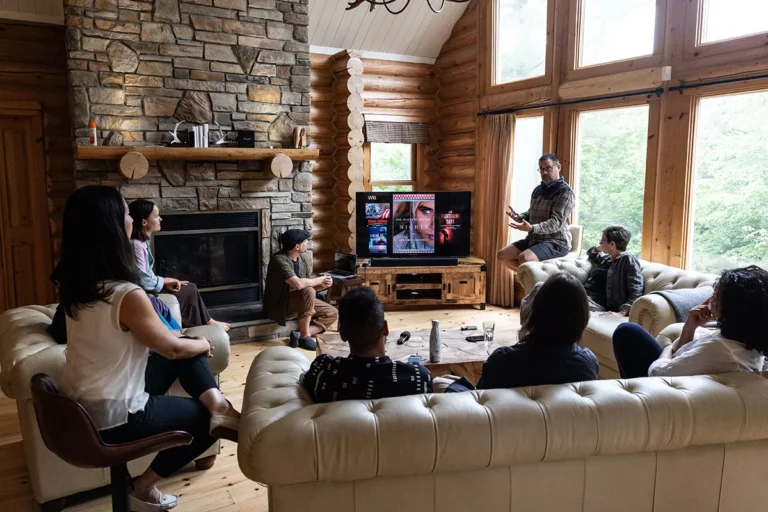 Seven people gather in a cozy log cabin living room with large windows, watching TV together. One stands near the TV, gesturing as if explaining something. The scene, captured by Rebels and Rascals Photography, features a stone fireplace and wooden walls.