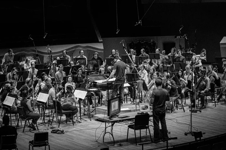 A black and white photo by Rebels and Rascals Photography captures an orchestra rehearsing on stage, with musicians playing various instruments and a conductor at the center. Microphones and equipment are set up around the group.