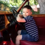 A young boy wearing a navy striped shirt and denim shorts sits in the driver’s seat of a vintage car, smiling and holding the steering wheel. The cars red interior with wood paneling is captured beautifully by Rebels and Rascals Photography.