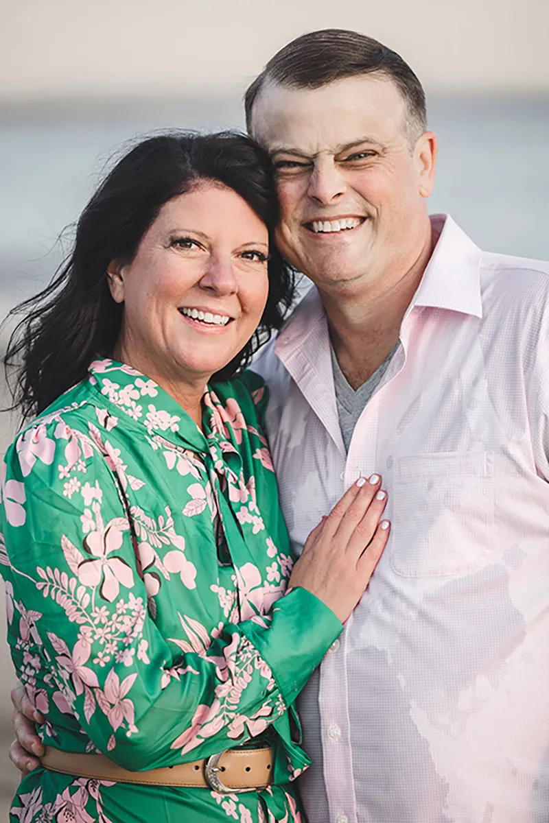 A smiling couple stands close together outdoors by Rebels and Rascals Photography. The woman wears a green floral dress, the man a light pink shirt; they look happy with dark and light hair, with the beach in the background.